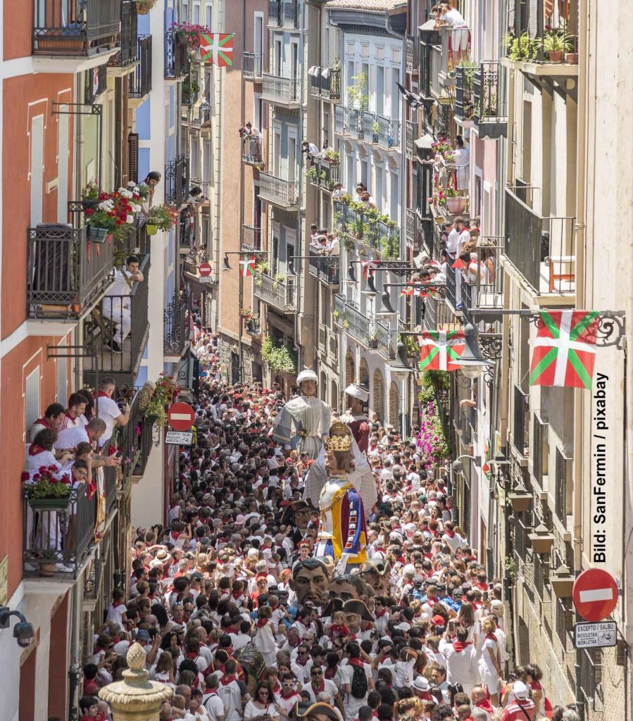 Eine überfüllte Gasse in einer Altstadt, gesäumt von mehrstöckigen Gebäuden mit Balkonen, die mit Blumen geschmückt sind. Eine Parade bewegt sich durch die Mitte, und große Figuren von Giganten, geschmückt mit Kleidung und Flaggen, ragen über die Menschenmenge hinaus. Die Gasse ist belebt und festlich.