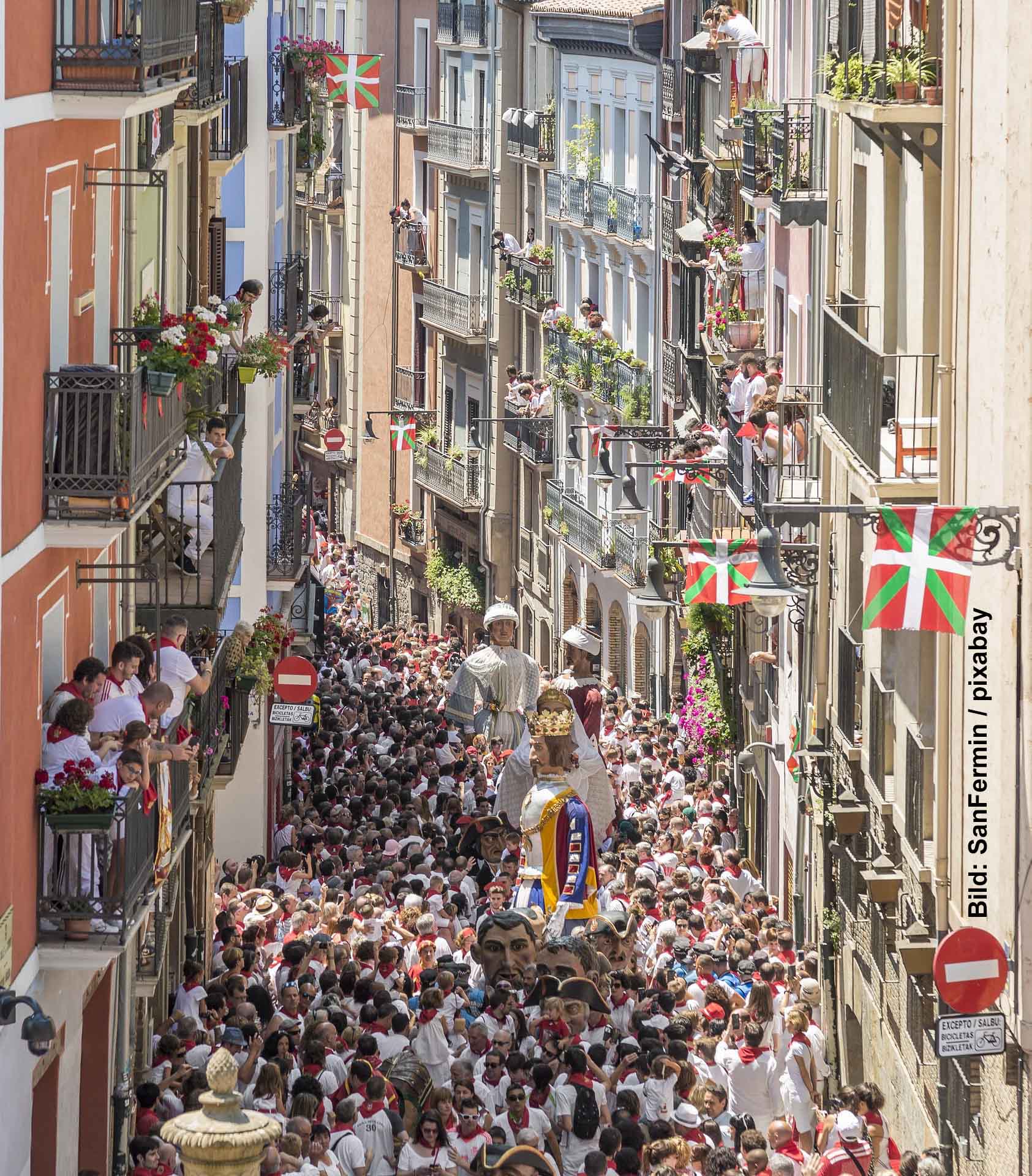 Eine überfüllte Gasse in einer Altstadt, gesäumt von mehrstöckigen Gebäuden mit Balkonen, die mit Blumen geschmückt sind. Eine Parade bewegt sich durch die Mitte, und große Figuren von Giganten, geschmückt mit Kleidung und Flaggen, ragen über die Menschenmenge hinaus. Die Gasse ist belebt und festlich.