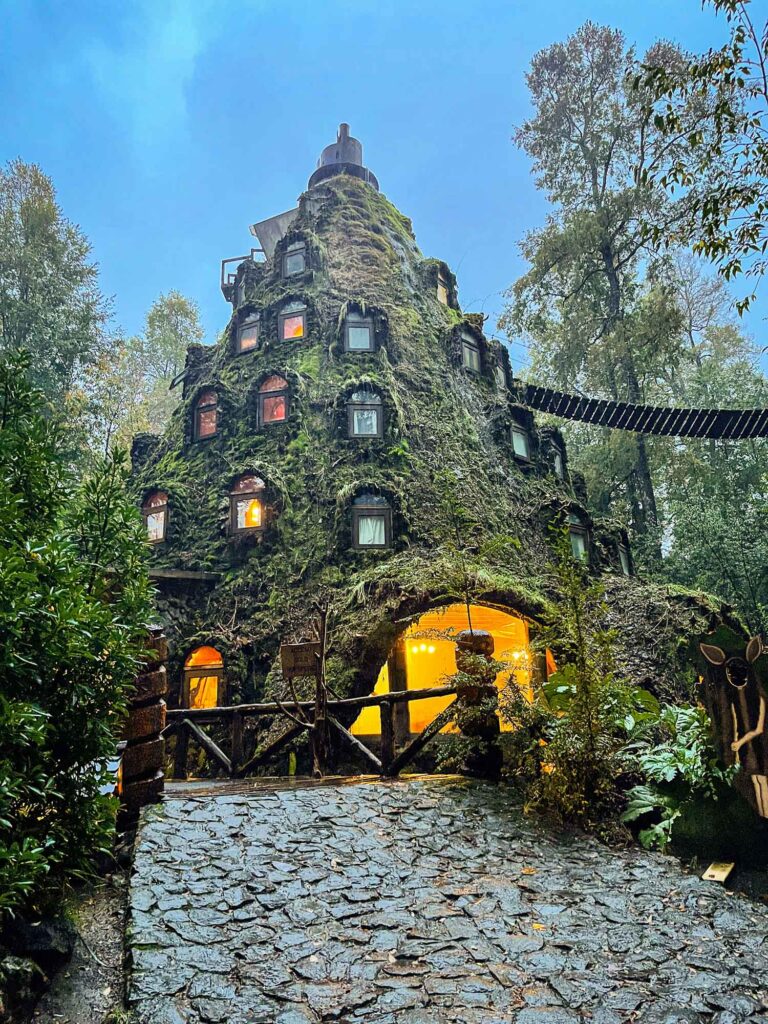 Huilo Huilo: Das magische Herz des chilenischen Regenwaldes 6 Eine Abendaufnahme des kegelförmigen, moosbewachsenen "Montana Magica" Hotels im dichten Wald. Die unregelmäßig angeordneten Fenster strahlen warmes, oranges Licht aus. Auch der Eingangsbereich ist hell beleuchtet. Ein gepflasterter Steinweg führt zum Gebäude. Oben rechts ist ein Teil einer Hängebrücke zu sehen. Der Himmel ist in tiefem Blau und Grau gehalten. Eine kleine Hirschfigur ist rechts im Gebüsch zu sehen.