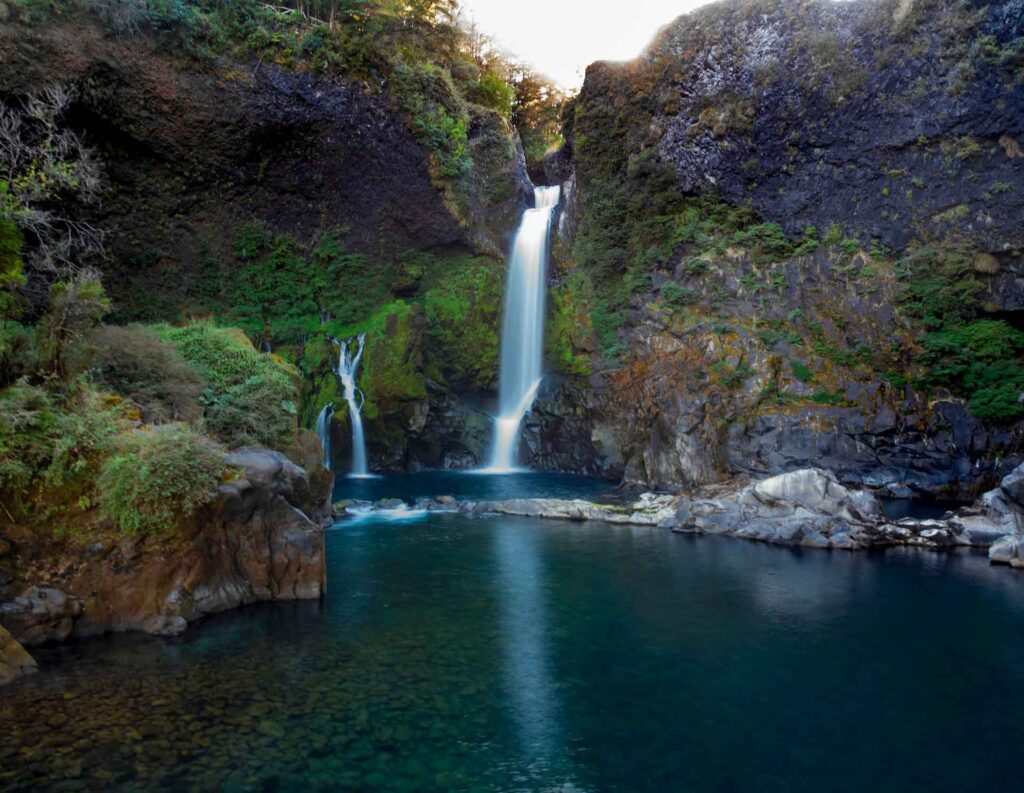 Huilo Huilo: Das magische Herz des chilenischen Regenwaldes 4 Ein Landschaftsfoto eines Wasserfalls, der über eine hohe, dunkle Felswand stürzt. Der Hauptwasserstrom fällt in ein klares, tiefblaues Wasserbecken. Ein kleinerer Wasserfall ist links daneben zu sehen. Die Felsen sind teilweise mit grünem Moos und Pflanzen bewachsen. Im Vordergrund ist das Wasser des Pools so klar, dass man die Steine auf dem Grund sehen kann. Am oberen Rand bricht Sonnenlicht durch die Bäume.