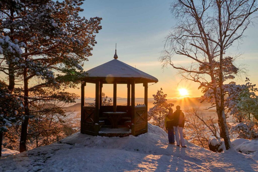 Zwei Wandernde stehen im Schnee neben einem hölzernen Aussichtspavillon auf dem Gohrisch und blicken in den Sonnenaufgang.