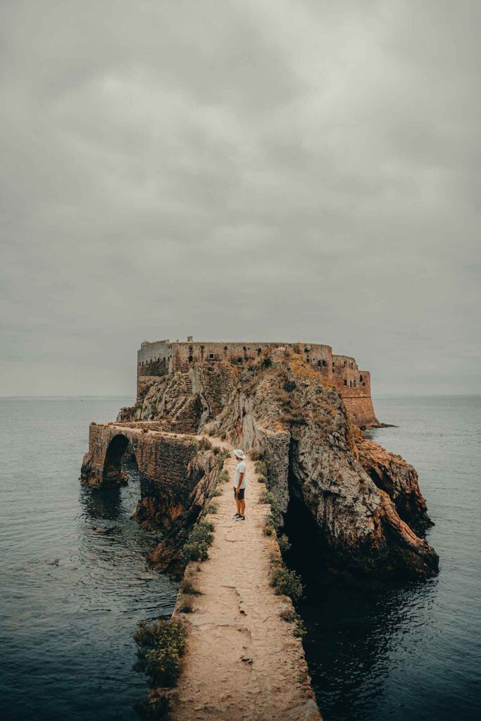 Person auf schmalem Steinweg zur Festung São João Baptista auf den Berlengas, links und rechts das Meer unter grauem Himmel.