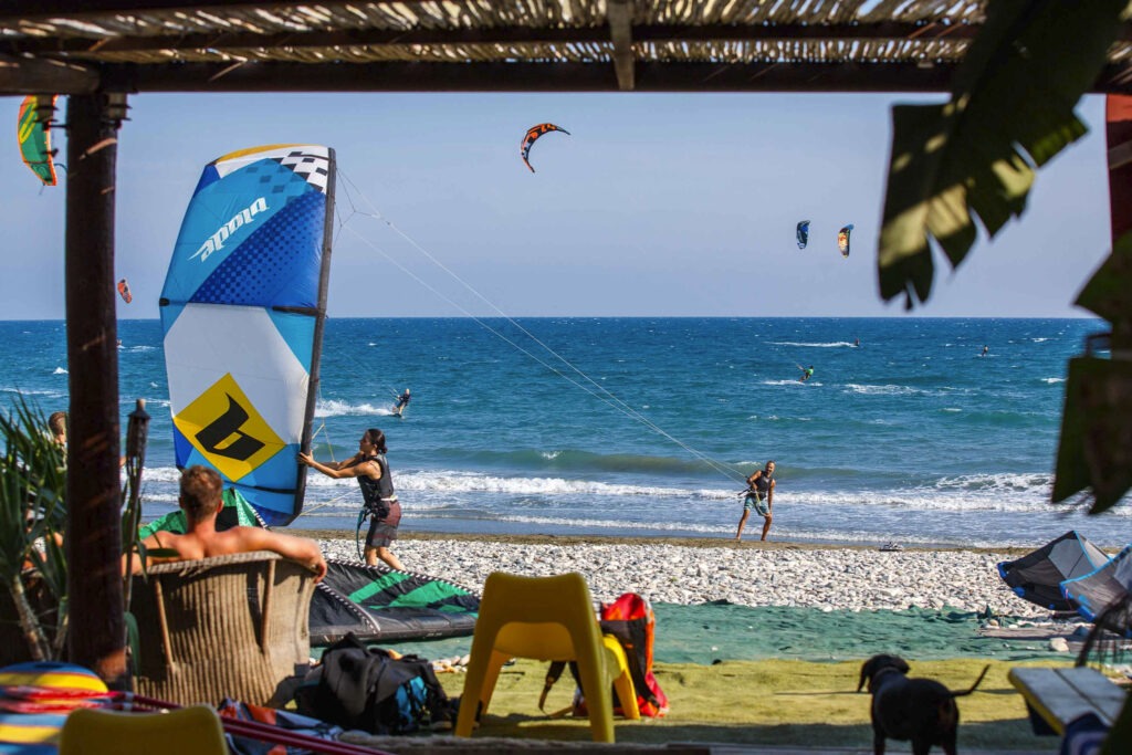 Mehrere Kitesurfer mit bunten Lenkdrachen auf dem Meer; im Vordergrund Strandgäste und Surfausrüstung.