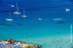 Türkisblaues Meer in der Konnos Bay mit Segelbooten, vielen Schwimmern und einem Strandabschnitt mit Sonnenschirmen.