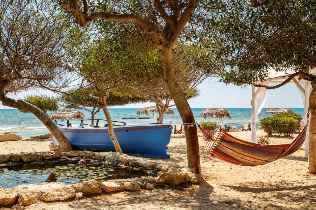 Blaues Boot und gestreifte Hängematte unter Bäumen an einem Sandstrand bei Agia Napa.