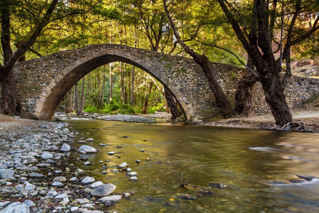 Alte Steinbogenbrücke über einem flachen Fluss zwischen Bäumen im Troodos-Gebirge.