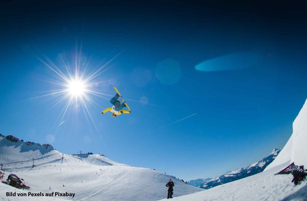 Ein Snowboarder vollführt einen spektakulären Sprung kopfüber vor einer strahlenden Sonne in einer verschneiten Berglandschaft.