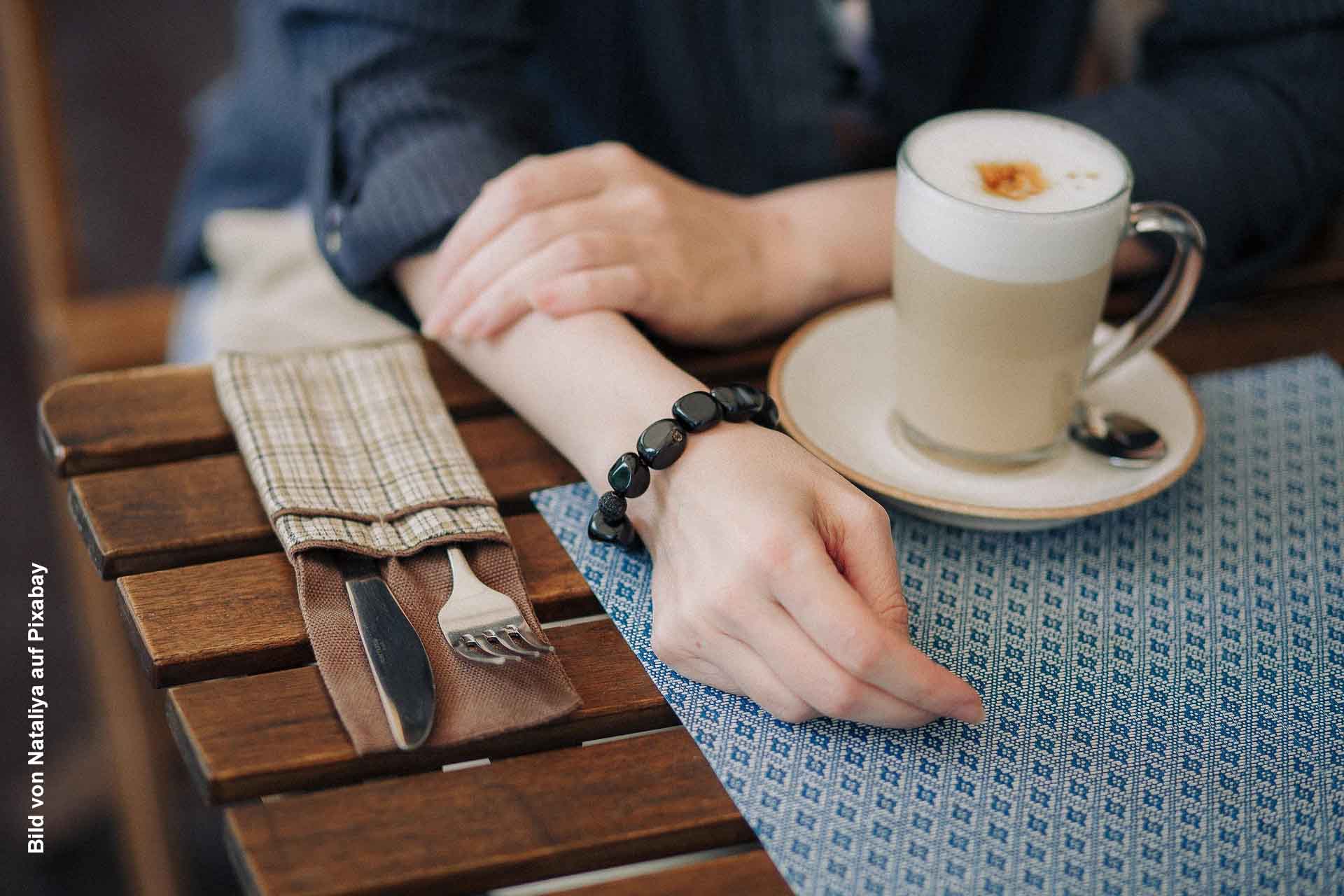 Eine Person sitzt an einem Holztisch im Café, vor ihr eine Tasse Cappuccino auf einer Untertasse, daneben Besteck in einer Stoffhülle und ein blau gemustertes Platzset.