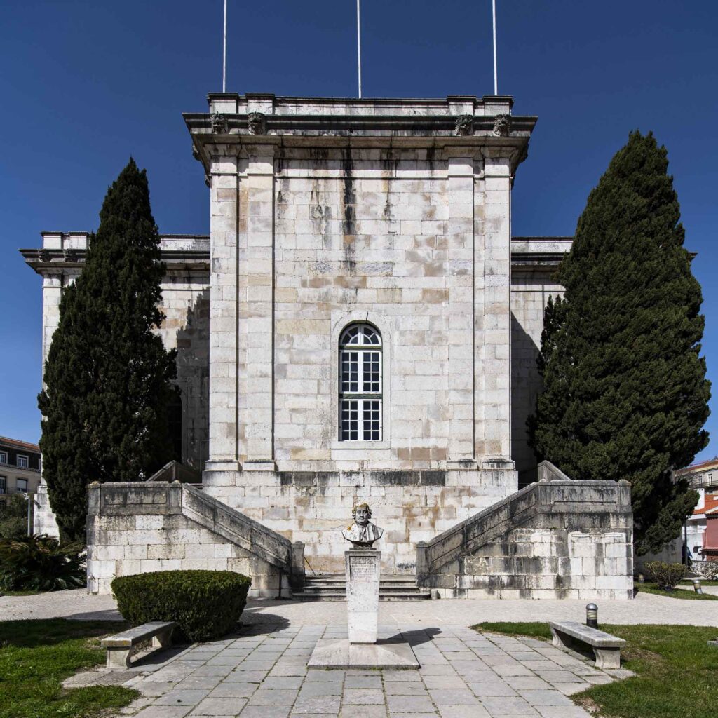 Außenansicht des historischen Wasserspeichers Mãe d’Água in Lissabon mit Steintreppe, Büste vor dem Gebäude und zwei hohen Zypressen.