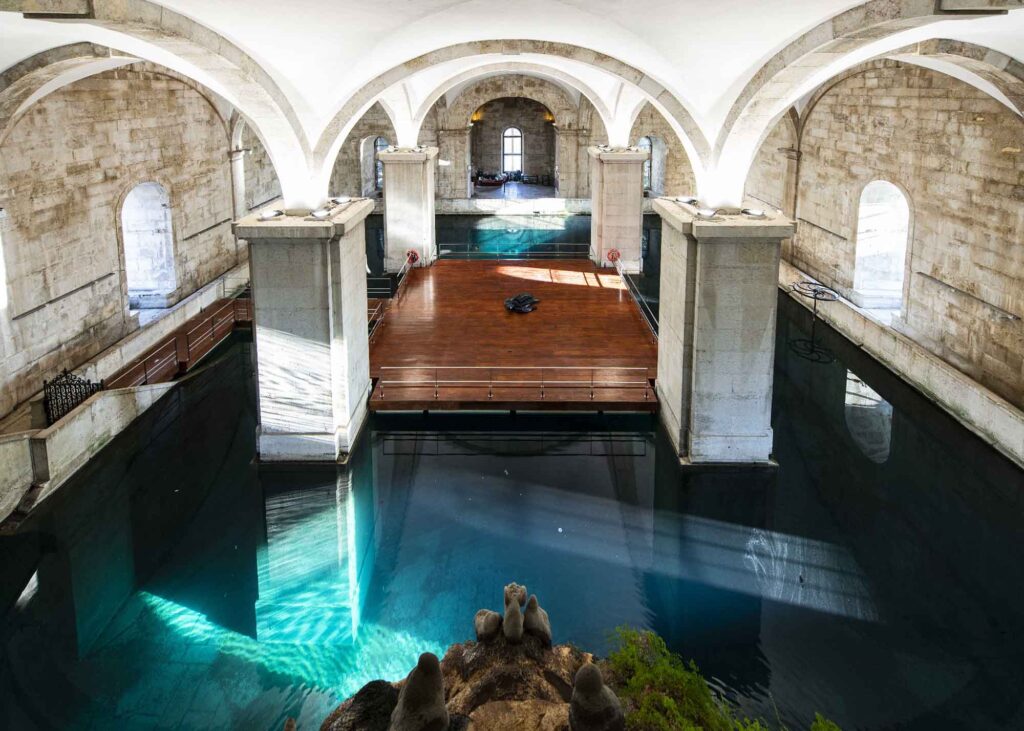 Historischer Wasserspeicher Mãe d’Água in Lissabon mit großen Steinpfeilern, weißen Gewölben, Wasserbecken und einer hölzernen Plattform.