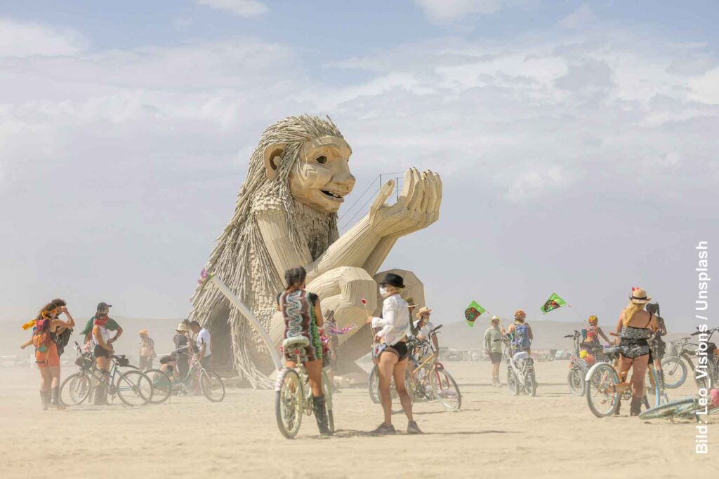 Große sitzende Holzfigur beim Burning Man, umgeben von Besucherinnen und Besuchern mit Fahrrädern auf der staubigen Playa.