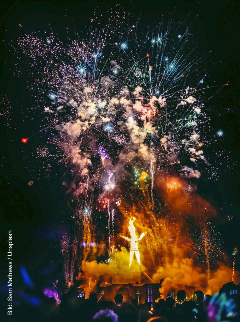 Brennende Burning-Man-Figur bei Nacht unter einem großen Feuerwerk, davor die Silhouetten zahlreicher Zuschauer.