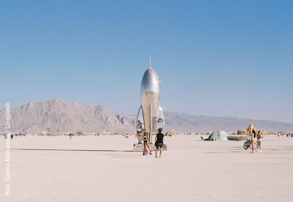 Raketenförmige Kunstinstallation beim Burning Man auf der Playa, mit Besuchern und Bergen im Hintergrund.