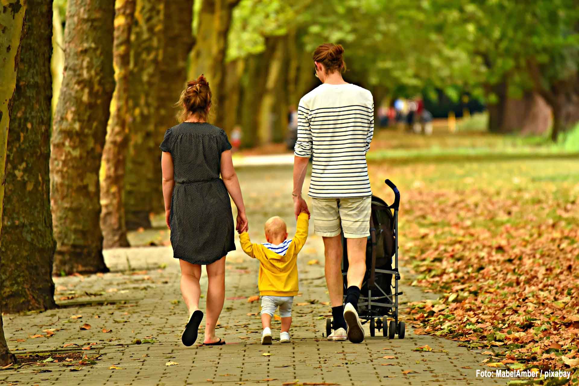 Ein Paar mit einem Kleinkind läuft uns den Rücken zugewandt durch einen Park