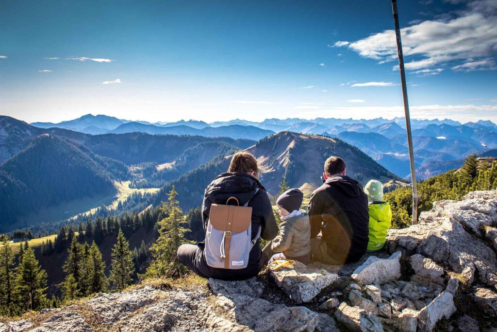 Reisetrend Mikroabenteuer: Genussmomente direkt vor der Haustür 3 Vierköpfige Familie sitz am Berghang und schaut aufs Tal