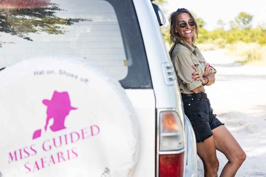 Wildnis erleben, Freiheit spüren - mit Miss Guided Safaris 3 Maika Kretschmer, Gründerin von Miss Guided Safaris, lehnt lachend an einem Geländewagen mit dem Firmenlogo im Hintergrund.