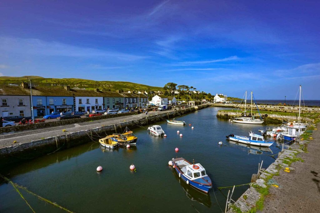 Blick auf den kleinen Hafen von Carnlough an der Nordküste Irlands mit bunten Fischerbooten, Pubs und grünen Hügeln im Hintergrund