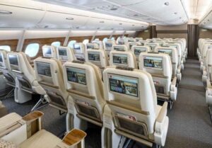 Emirates A380 Sitzplätze und Services - die Fotos 30 Blick von hinten auf mehrere Sitzreihen der Premium Economy Class. In den Rückseiten der hellen Vordersitze sind große Flachbildschirme integriert, die das Emirates-Unterhaltungsmenü zeigen. Unten sind Sitztaschen und Fußstützen zu sehen.