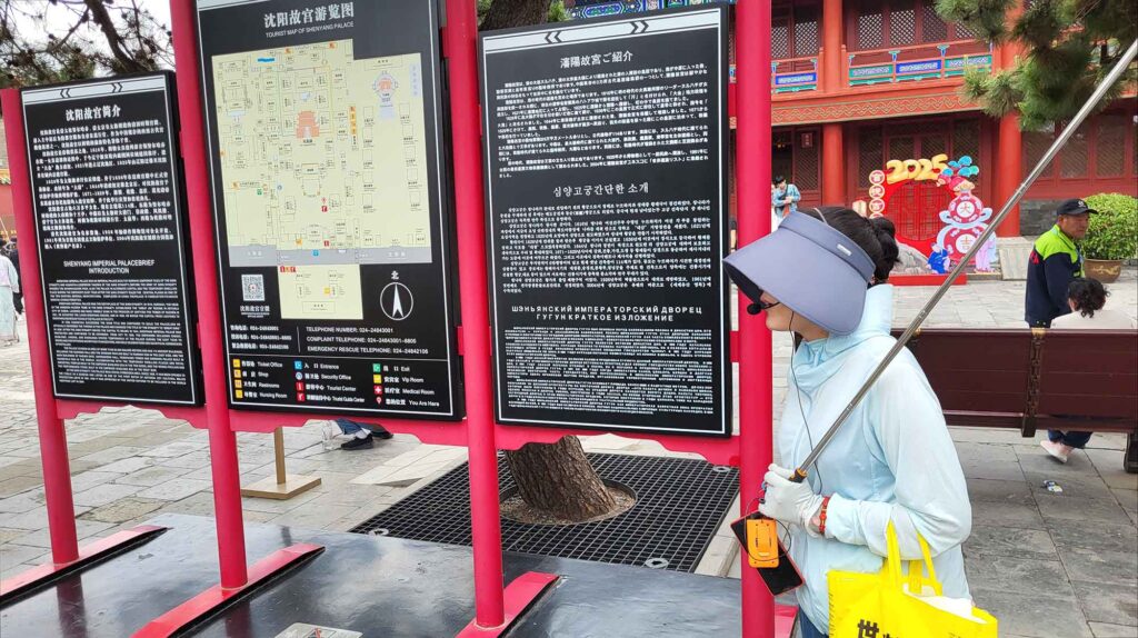Kaiserpalast von Shenyang – Die „Verbotene Stadt“ des Nordens erleben 21 Touristenführerin mit Visier vor schwarzer Informationstafel mit Lageplan und Texten (Chinesisch, Englisch, Russisch) am Eingang des Kaiserpalasts Shenyang.