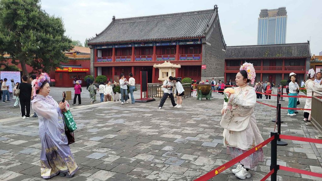 Kaiserpalast von Shenyang – Die „Verbotene Stadt“ des Nordens erleben 5 Zwei Frauen in Qing-Kaiserinnen-Kostümen fotografieren sich im Hof des Shenyang-Palasts; rote Wände, graue Ziegeldächer, weitere Touristen.