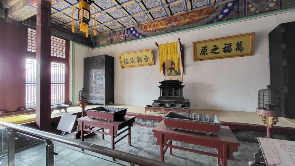 Kaiserpalast von Shenyang – Die „Verbotene Stadt“ des Nordens erleben 9 Der Innenraum eines Palastgebäudes mit zwei großen Schriftrollen an der Wand, eine große schwarze Holztruhe, einem Thronsessel auf einer erhöhten Plattform und einer bunt bemalten Decke mit Mustern.