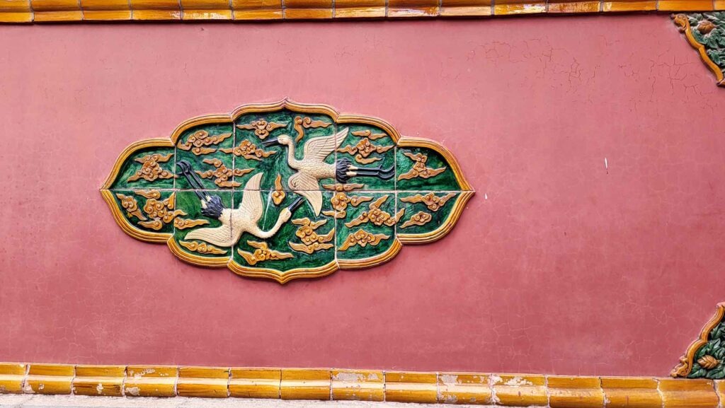 Kaiserpalast von Shenyang – Die „Verbotene Stadt“ des Nordens erleben 18 Nahaufnahme einer Wanddekoration im Shenyang-Kaiserpalast. Die Verzierung zeigt zwei goldene Kraniche, die in einem grünen, wolkenartigen Rahmen fliegen, der auf einer roten Wand angebracht ist.