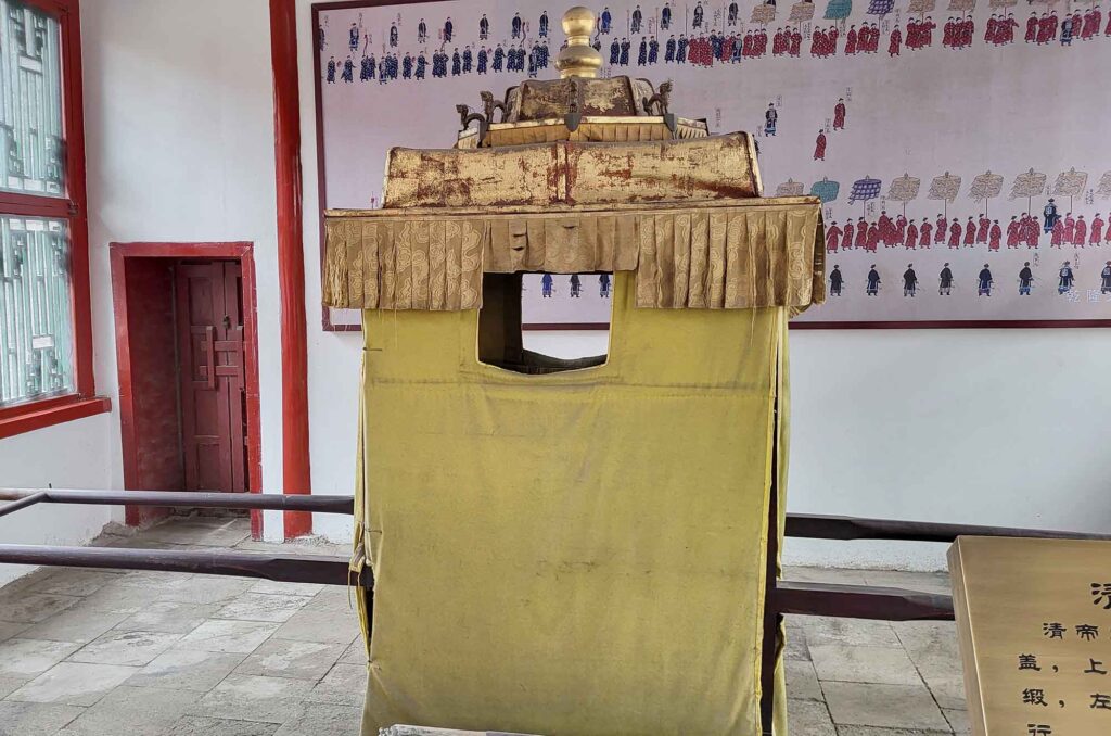 Kaiserpalast von Shenyang – Die „Verbotene Stadt“ des Nordens erleben 12 Eine antike gelbe Sänfte mit einem goldenen Dach und roten Tragebalken ist in einem Innenraum des Palastes ausgestellt. Im Hintergrund ist ein großer Wandteppich mit einer Darstellung von Personen in traditionellen Gewändern zu sehen.