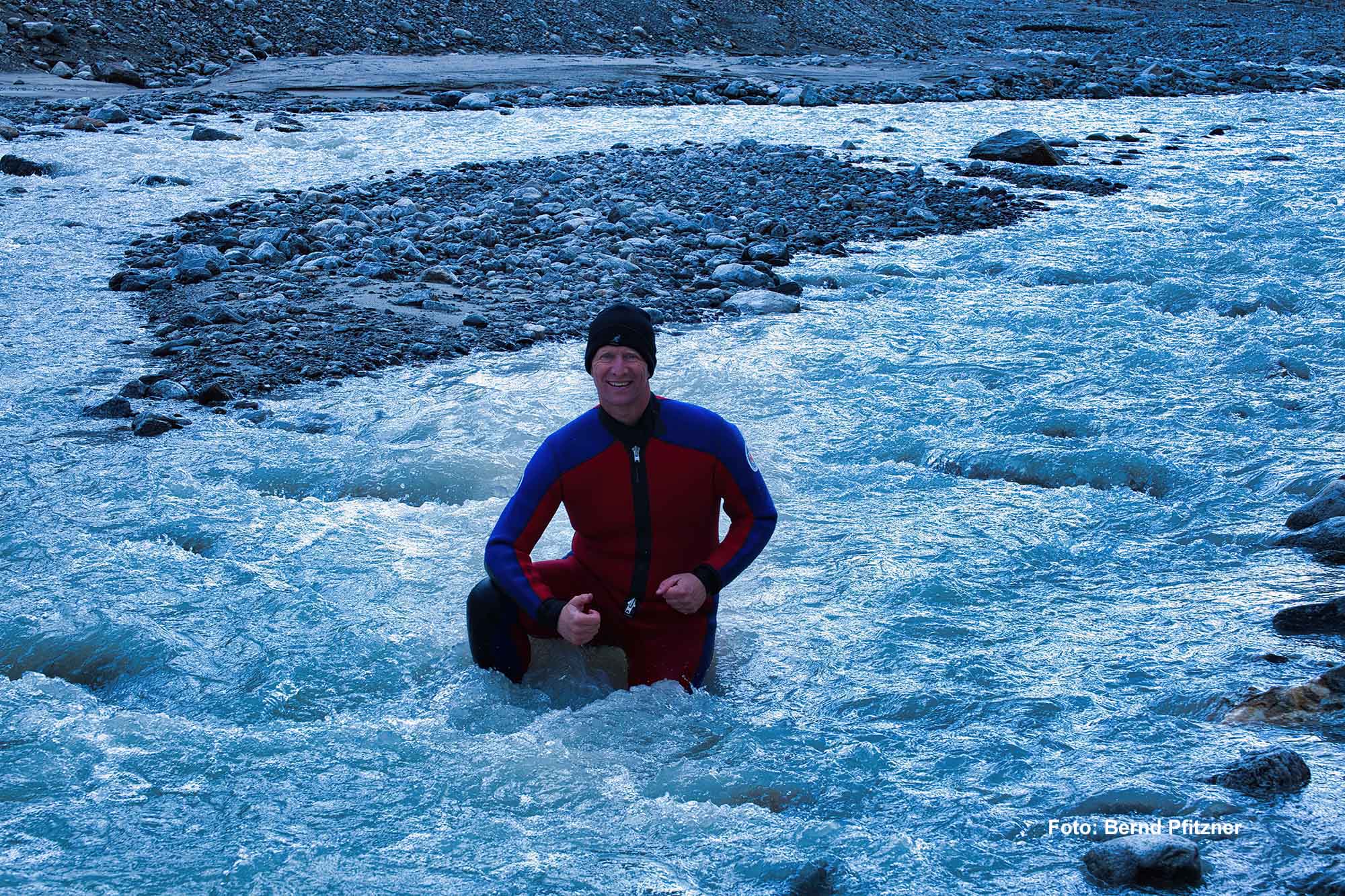 Mann im Neoprenanzug kniet lachend im eiskalten Gletscherwasser der Aarequelle.