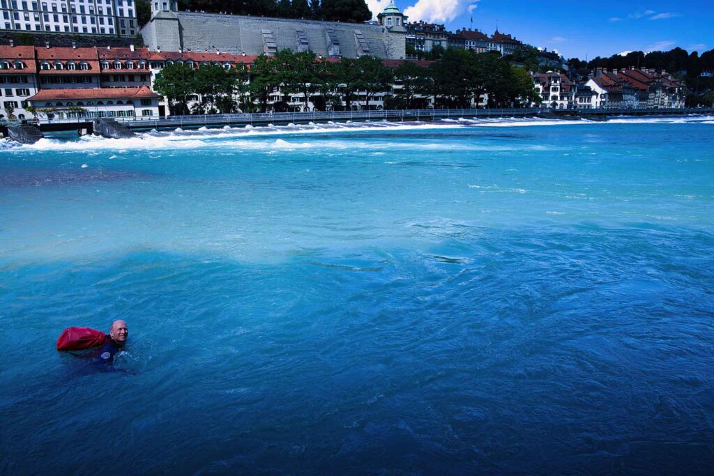 Durch eiskaltes Wasser und Stromschnellen schwimmen 5 Schwimmer mit Seesack im blauen Wasser der Aare vor Häuserkulisse in Bern
