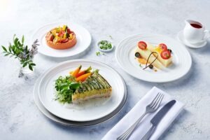 **Alt-Text:**
Veganes Drei-Gänge-Menü bei Emirates: kunstvoll angerichteter pflanzlicher Hauptgang mit geschichtetem Gemüse und Kräutern, dazu Vorspeise und veganes Dessert auf weißem Porzellan, serviert in stilvoller Airline-Präsentation.
