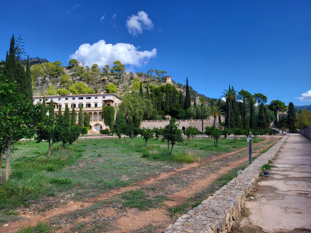 Blick auf ein historisches Herrenhaus mit Zitronenhain, Palmen und Zypressen in Sóller auf Mallorca, eingerahmt von Bergen und blauem Himmel.