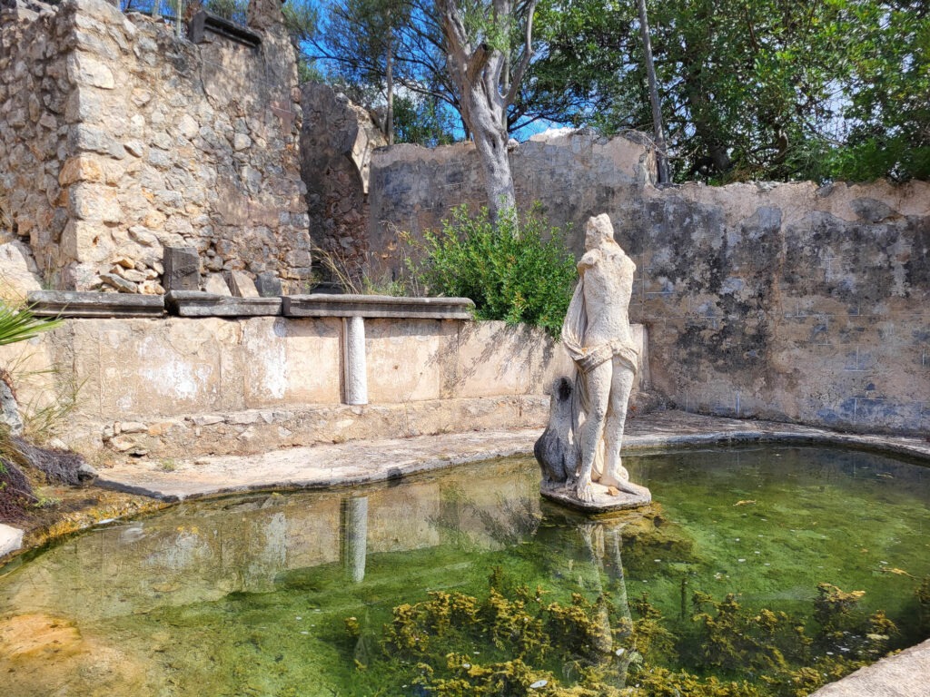 Historischer Brunnen mit Steinfigur in den Gärten des Landguts Raixa auf Mallorca, umgeben von alten Mauern.