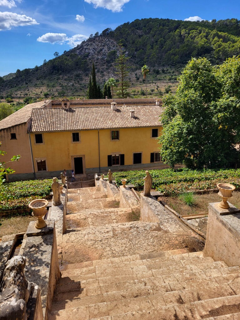Steintreppe mit Figuren und Blumenschalen im Landgut Raixa auf Mallorca, mit Blick auf das Herrenhaus und die Serra de Tramuntana.