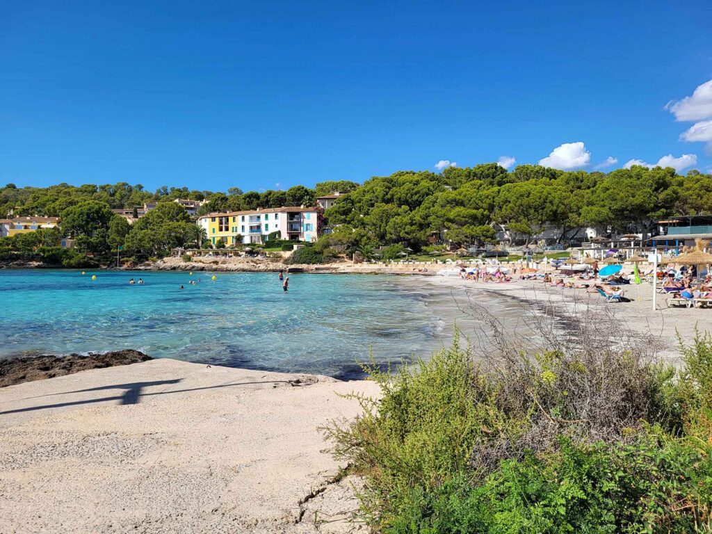 Playa Illetas – Mallorcas kleine Traumbuchten 4 Panorama des Strandes von Playa Illetas auf Mallorca mit türkisfarbenem Wasser, Badegästen und bunten Häusern am Ufer.