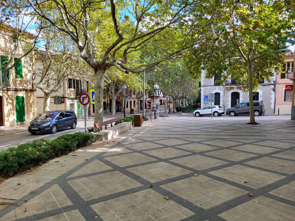 Ruhiger Platz in Esporles auf Mallorca mit gepflastertem Boden, alten Bäumen und Häusern mit grünen Fensterläden.