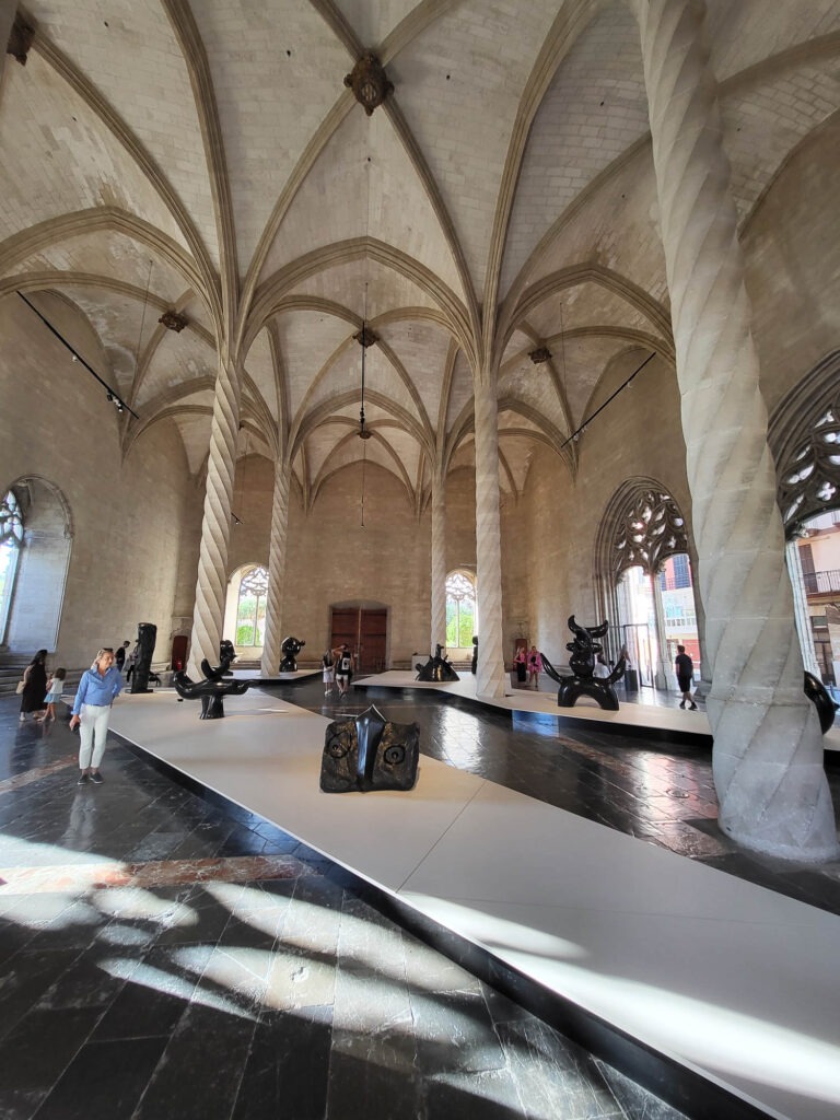 Das Foto zeigt einen beeindruckenden gotischen Saal mit hohen Kreuzrippengewölben und gedrehten Säulen, in dem moderne Skulpturen ausgestellt sind.