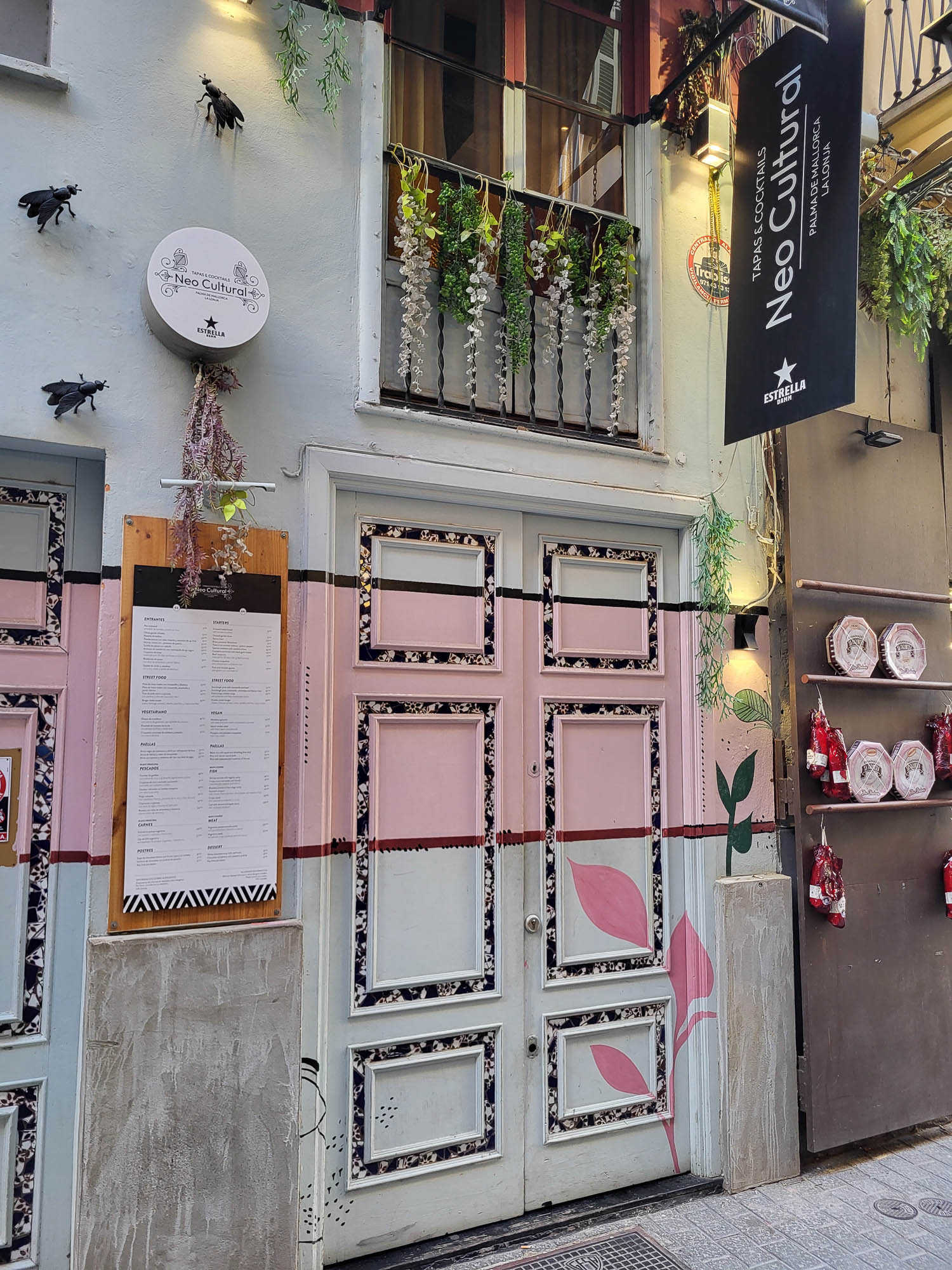 Auf dem Foto ist die bunt gestaltete Eingangstür eines Restaurants in einer schmalen Gasse zu sehen. An der Fassade hängen Deko-Elemente wie übergroße schwarze Fliegen, Pflanzen und ein Schild.