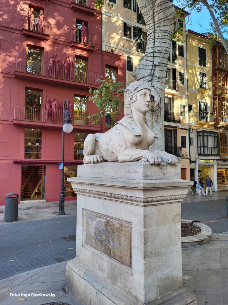 Das Foto zeigt eine Steinsphinx auf einem Sockel, im Hintergrund die farbigen Fassaden des Passeig del Born in Palma de Mallorca.