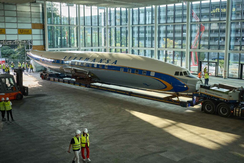 Zwei Lufthansa-Legenden vereint: Ju 52 und Lockheed Super Star in einer Ausstellung 4 Der restaurierte Rumpf einer Lufthansa Lockheed Super Star wird auf einem Schwertransporter in den Hangar One am Frankfurter Flughafen gezogen, während Mitarbeitende in Warnwesten den Vorgang begleiten.
