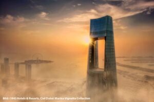 Atemberaubende Luftaufnahme des Ciel Dubai Marina bei Sonnenuntergang, mit goldenem Licht hinter dem markanten, hoch aufragenden Hotelturm und Blick auf die Skyline und Küste Dubais.