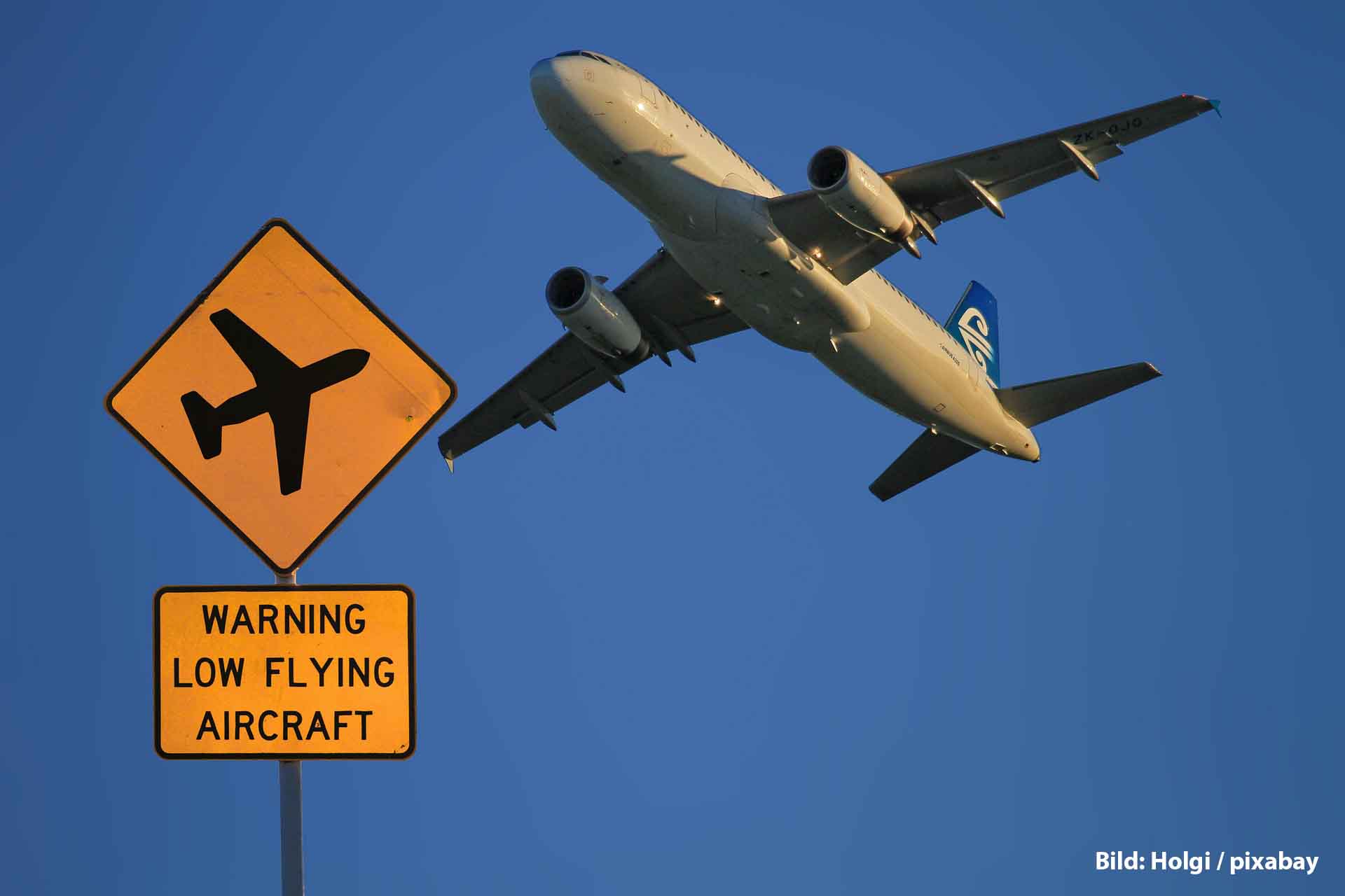 Airbus A320: Warum aktuell viele Maschinen ein Software-Update brauchen 1 Ein Airbus A320 fliegt tief über ein gelbes Warnschild mit der Aufschrift „Warning Low Flying Aircraft“ vor blauem Himmel.