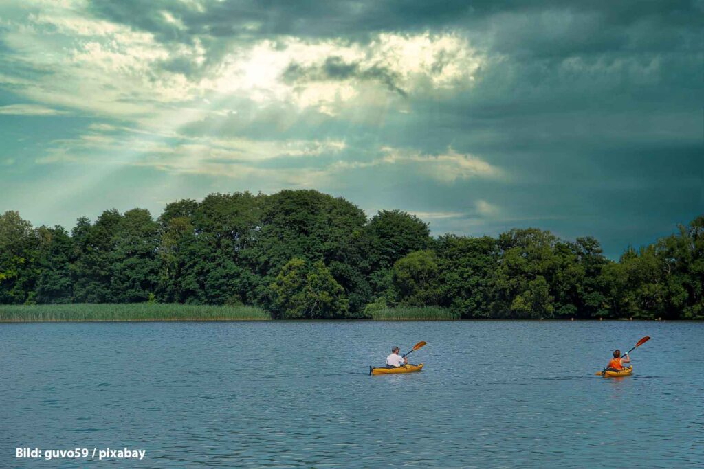 Zwei Menschen paddeln in gelben Kajaks über einen stillen See, im Hintergrund dichter Uferwald und dramatische Wolken mit Sonnenstrahlen.