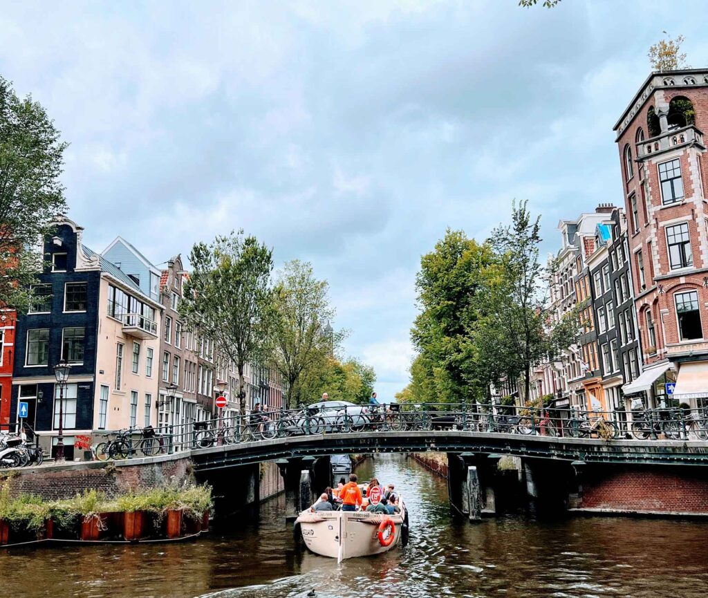 Städte vom Wasser aus entdecken: Eine neue Perspektive auf Europas Metropolen 3 Kleines Ausflugsboot fährt unter einer historischen Steinbrücke durch eine Amsterdamer Gracht, umgeben von Bäumen und alten Häusern.