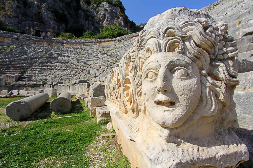 Detailaufnahme einer kunstvoll gemeißelten Steinskulptur im antiken Theater von Myra, Türkei; im Hintergrund halbkreisförmige Sitzreihen und verstreute Säulenfragmente.