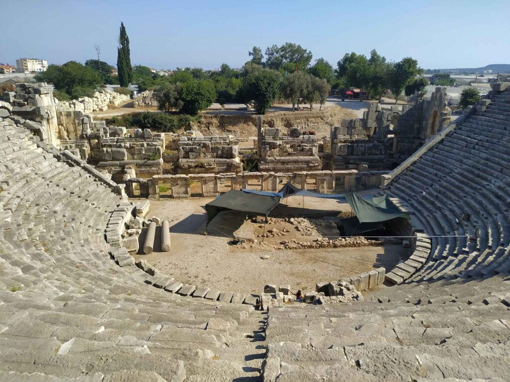 Blick in das antike Theater von Myra in der Türkei mit steinernen Sitzreihen, ausgegrabener Bühnenanlage und schattenspendenden Zelten im Zentrum; im Hintergrund Bäume und moderne Gebäude.