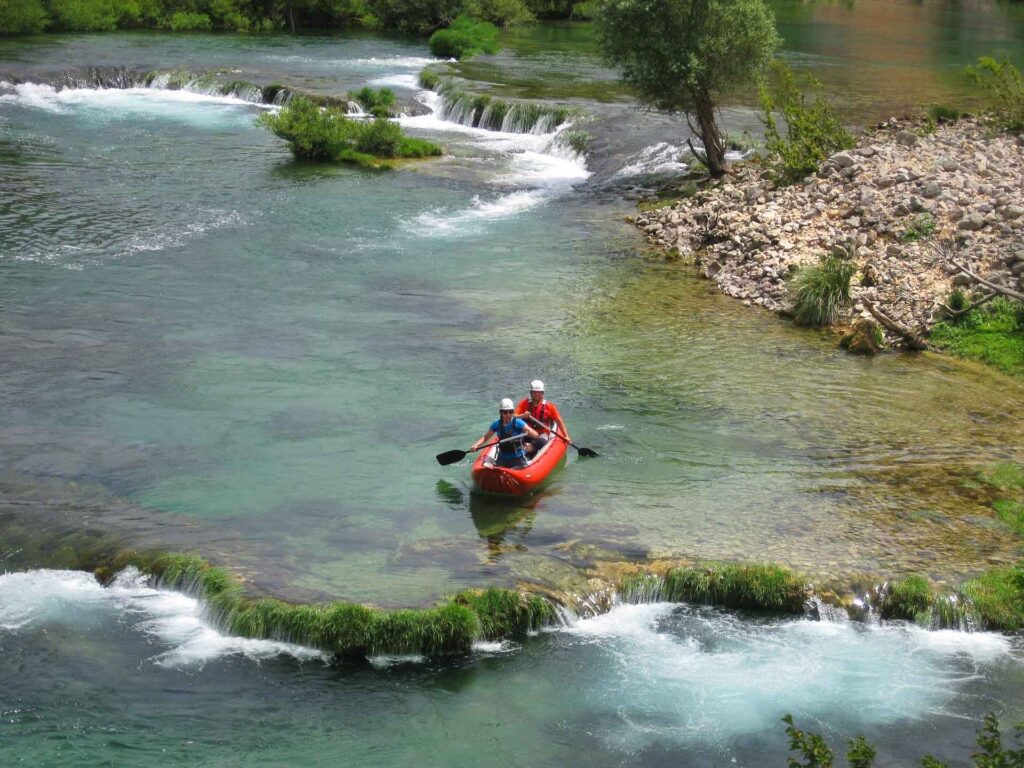 Zwei Personen in einem roten Kajak paddeln durch einen klaren Fluss mit flachen Stromschnellen, umgeben von grüner Ufervegetation und Felsen.