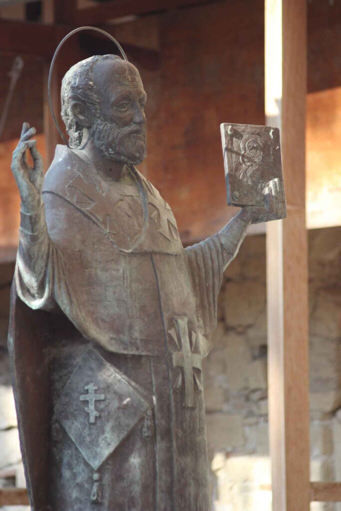 Bronzestatue des heiligen Nikolaus in liturgischer Kleidung, mit erhobener Hand und einer Ikone in der anderen; im Hintergrund eine steinerne Wand und Holzkonstruktion.