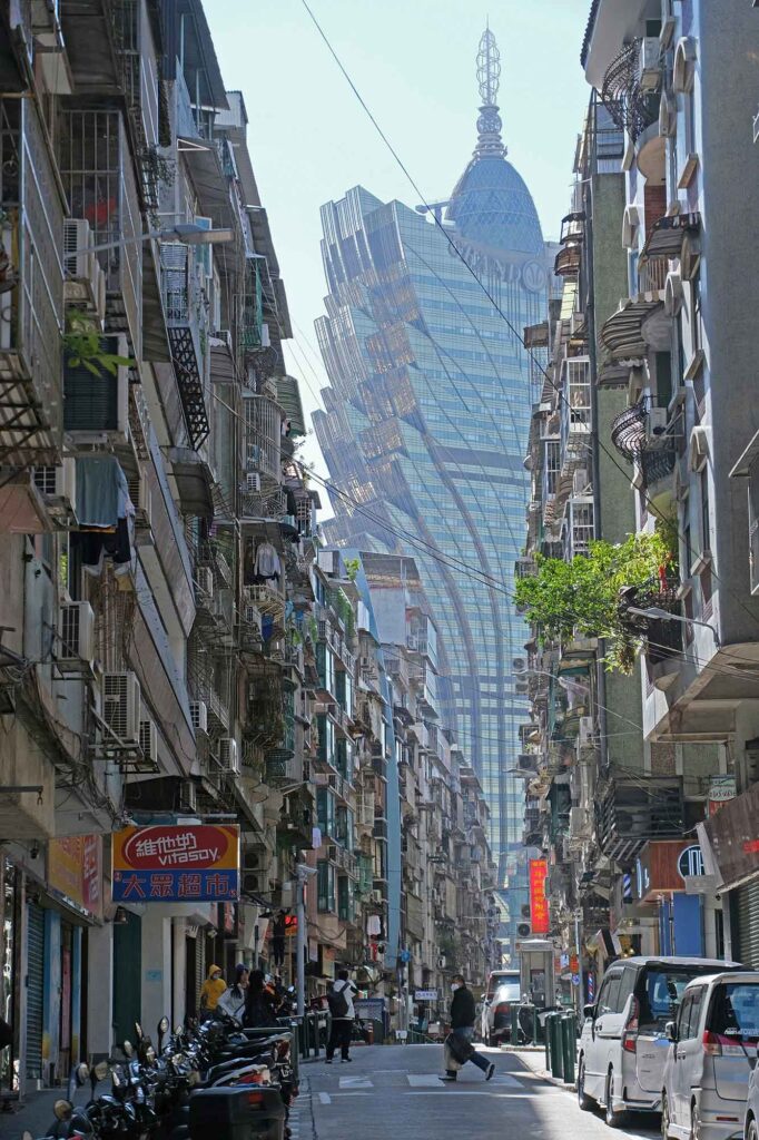 Blick durch eine enge Straße in Macau auf das auffällige Grand Lisboa Casino, eingerahmt von dicht bebauten Wohnhäusern und Straßengeschehen im Vordergrund.