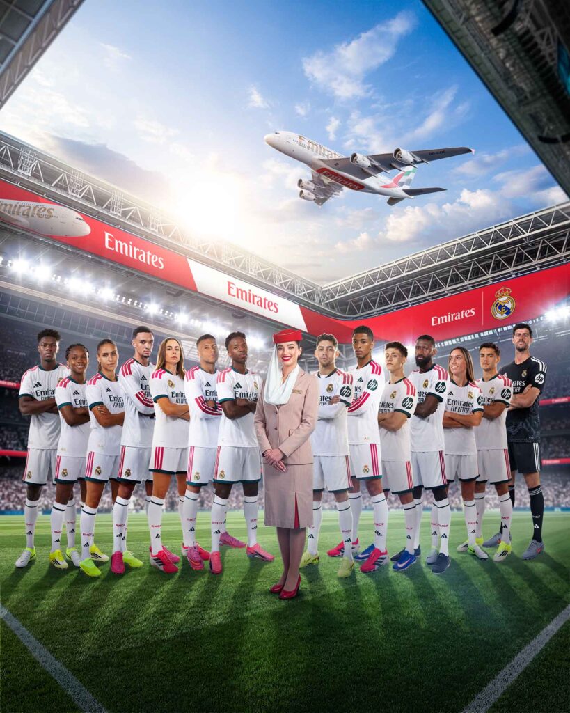 Eine Emirates-Kabinenmitarbeiterin steht gemeinsam mit Spielerinnen und Spielern von Real Madrid in einem Fußballstadion. Im Hintergrund sind Emirates-Bandenwerbung, das Real-Madrid-Wappen sowie ein startender Emirates Airbus A380 zu sehen. Das Motiv inszeniert die verlängerte Partnerschaft zwischen der Fluggesellschaft Emirates und dem spanischen Fußballclub Real Madrid.