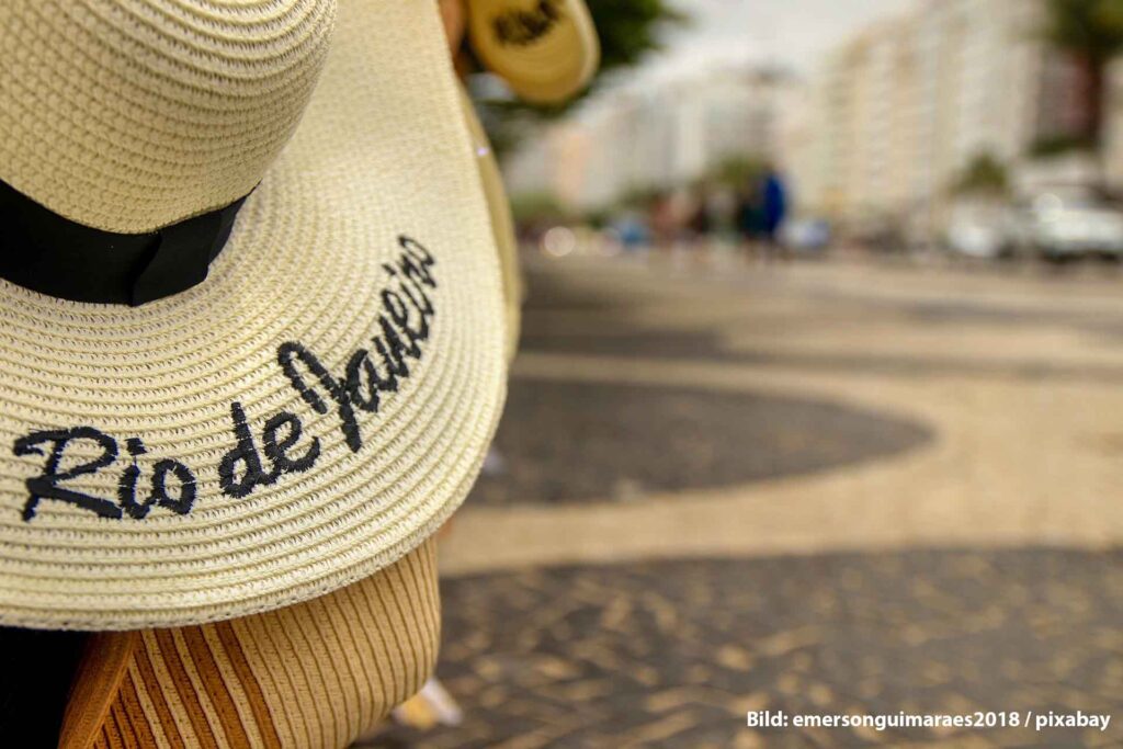 Start 1 Strohsonnenhut mit der schwarzen Aufschrift „Rio de Janeiro“ im Vordergrund, aufgenommen auf der berühmten wellenförmigen Pflasterpromenade von Copacabana; im Hintergrund verschwommene Häuser und Passanten.